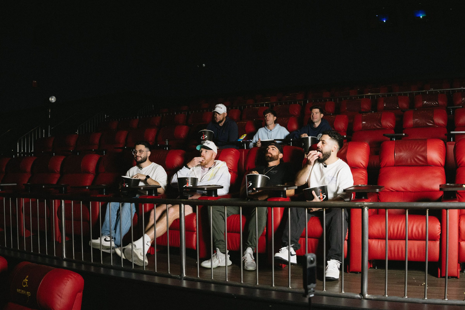 An audience seated in red recliner seats in a darkened cinema, watching the screen with popcorn.