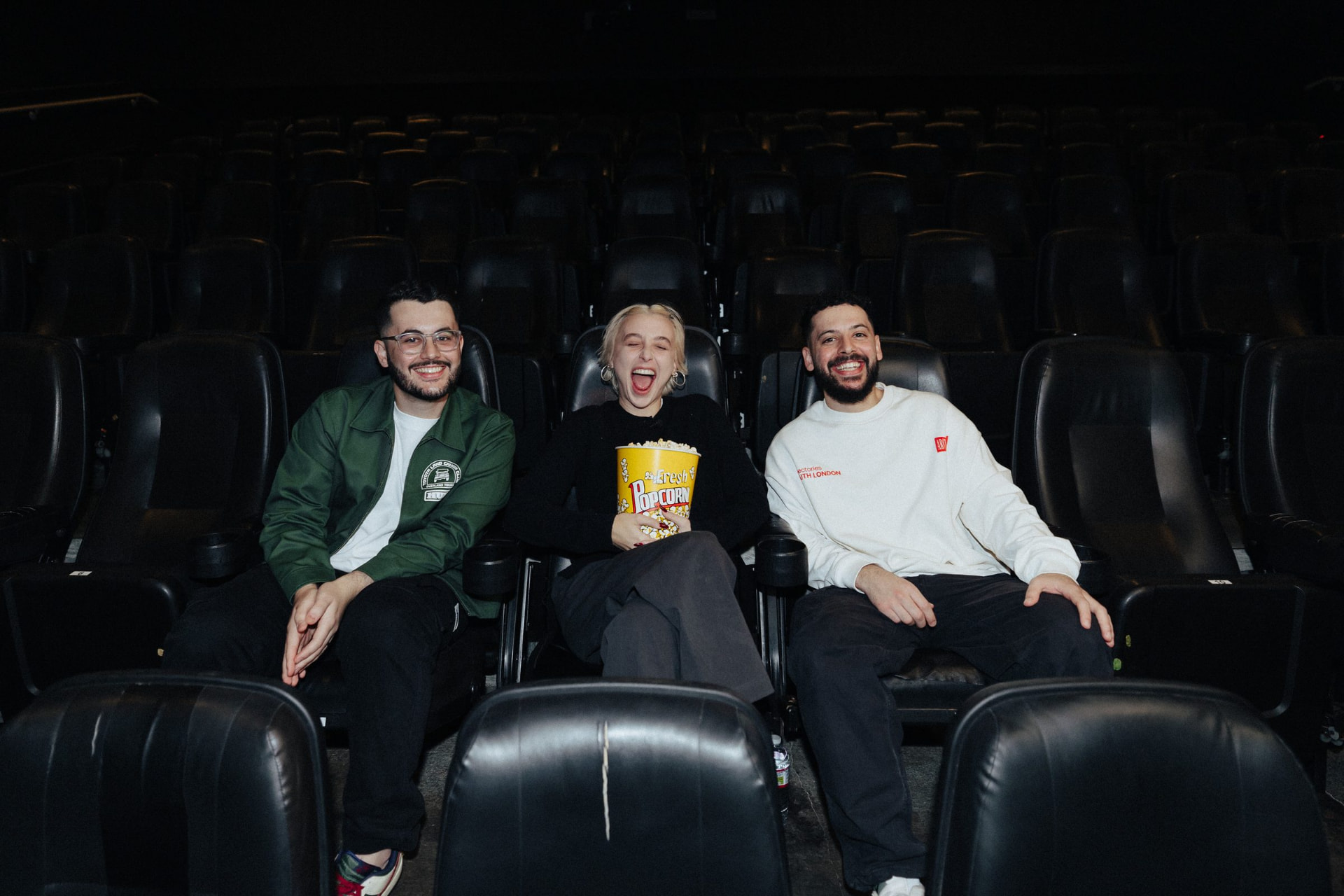 Three people sitting together in an empty auditorium, laughing while holding a tub of popcorn.