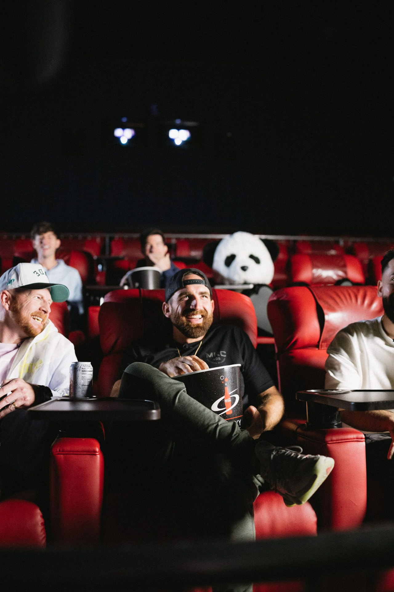 Vertical view of an audience in red cinema seats lit by the screen, projector beams visible above them.