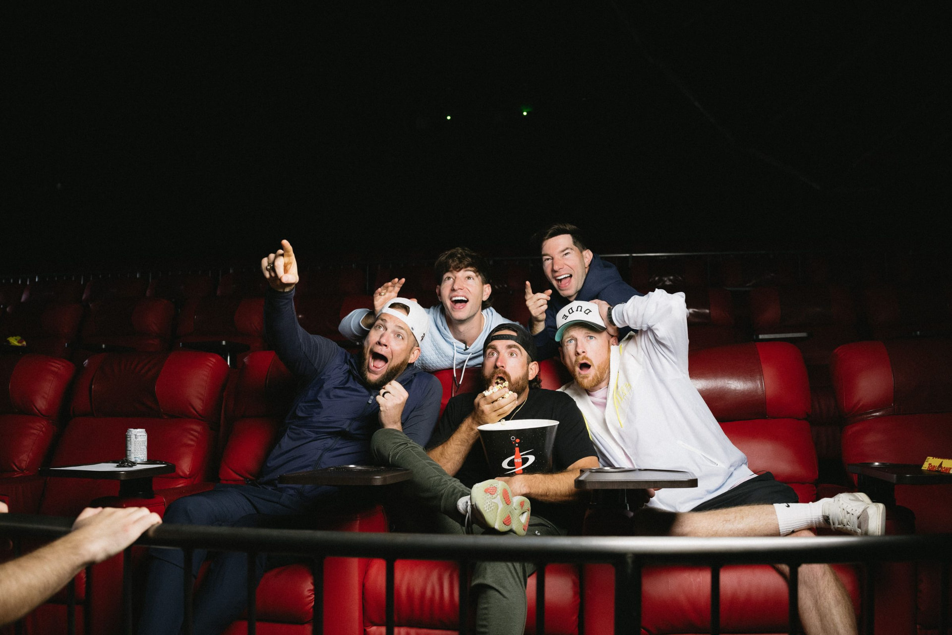 Five people in cinema seats reacting to the screen, one pointing and shouting.